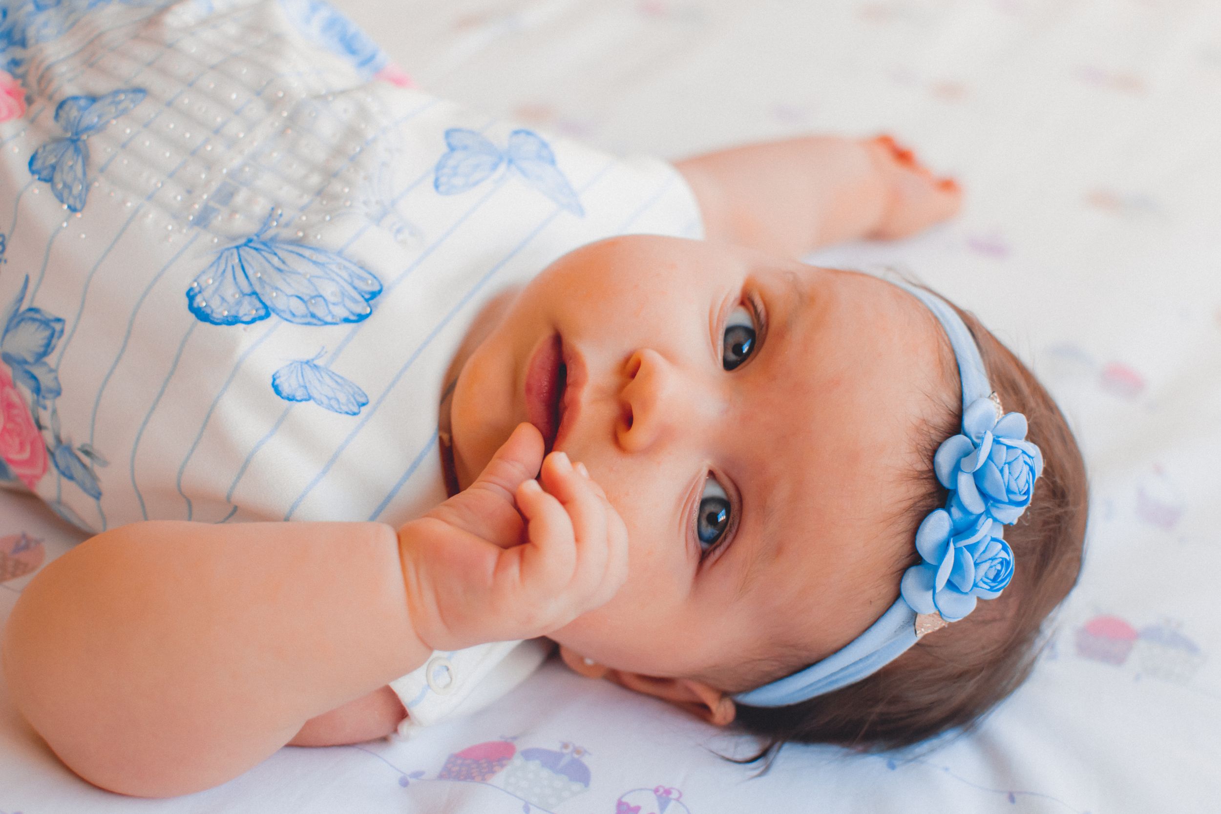 Baby with bright blue eyes wearing a delicate floral headband laying on patterned bedding.