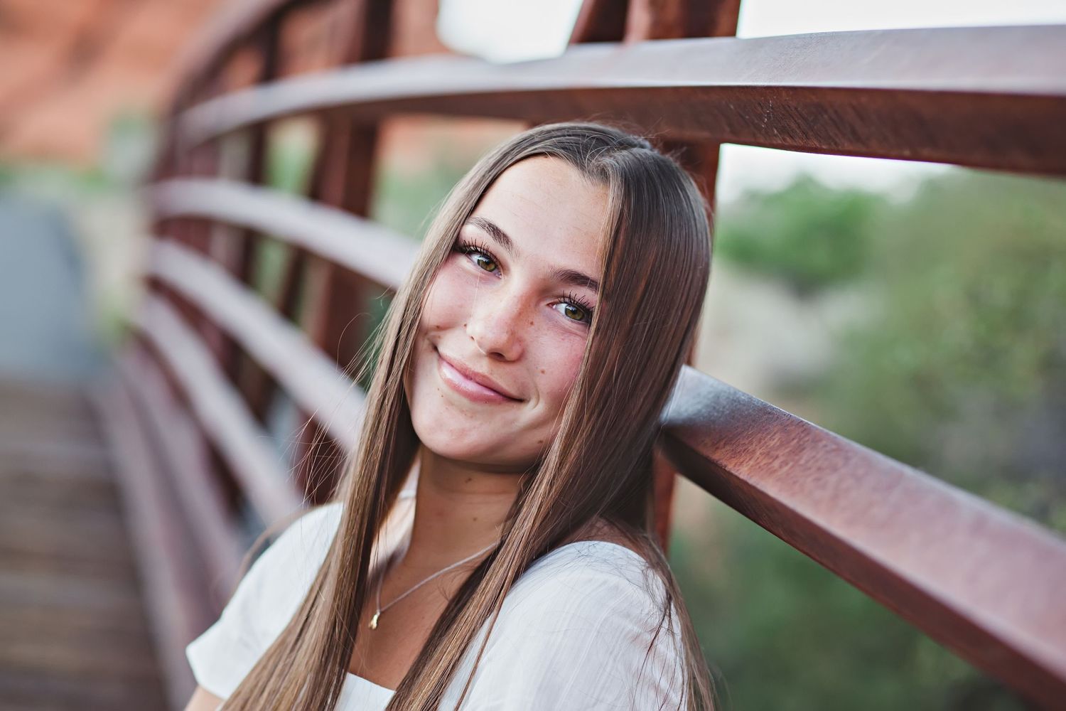 Portrait of someone with long brown hair smiling by a rustic bridge railing.