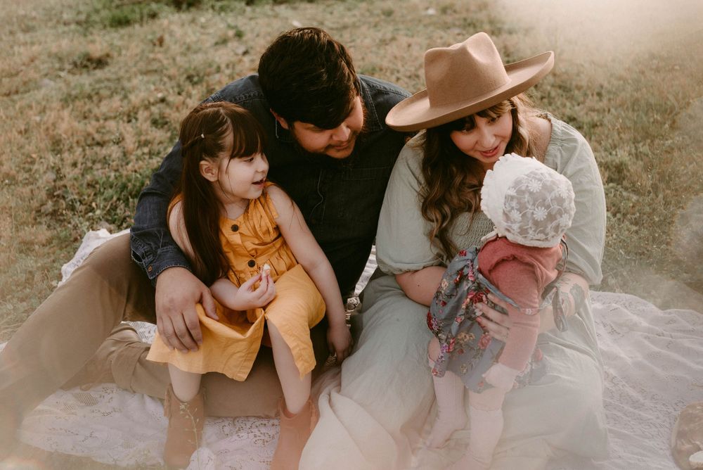 Intimate family portrait of parents and children sharing tender moment on picnic blanket at sunset.
