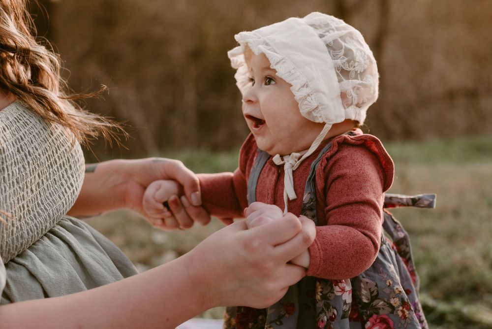 Young child in vintage bonnet and dress reaching towards someone during outdoor photoshoot.