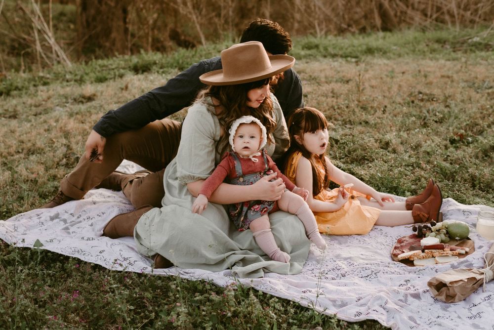 A cozy family picnic scene on a white lace blanket in a grassy field at sunset.
