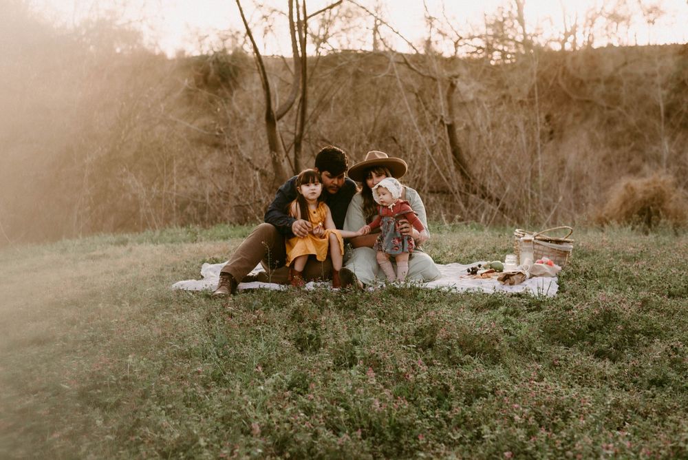 Family enjoying sunset picnic on grassy hillside with blanket and basket.