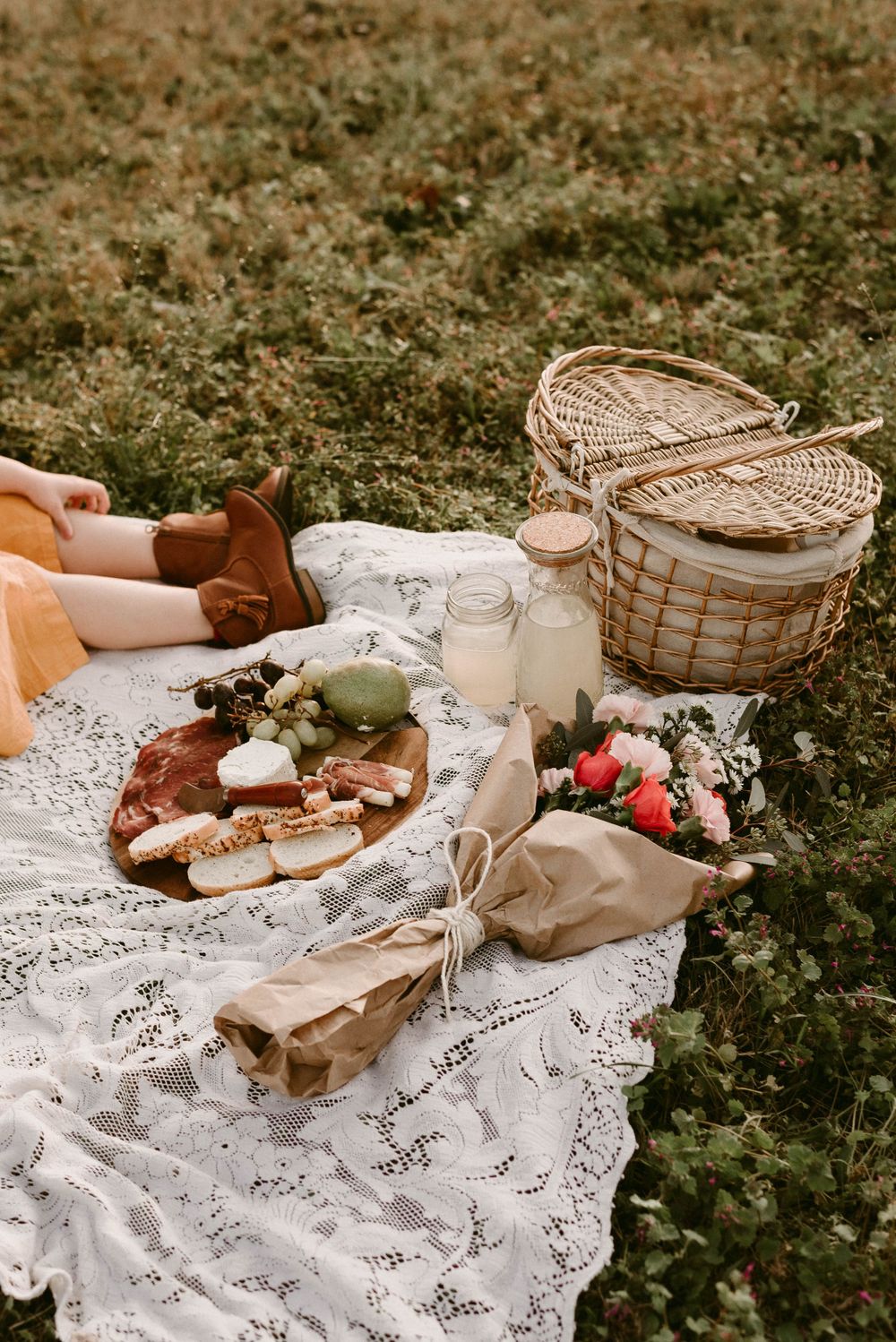 A rustic outdoor picnic setup with a wicker basket, charcuterie board, and lace blanket on grass.