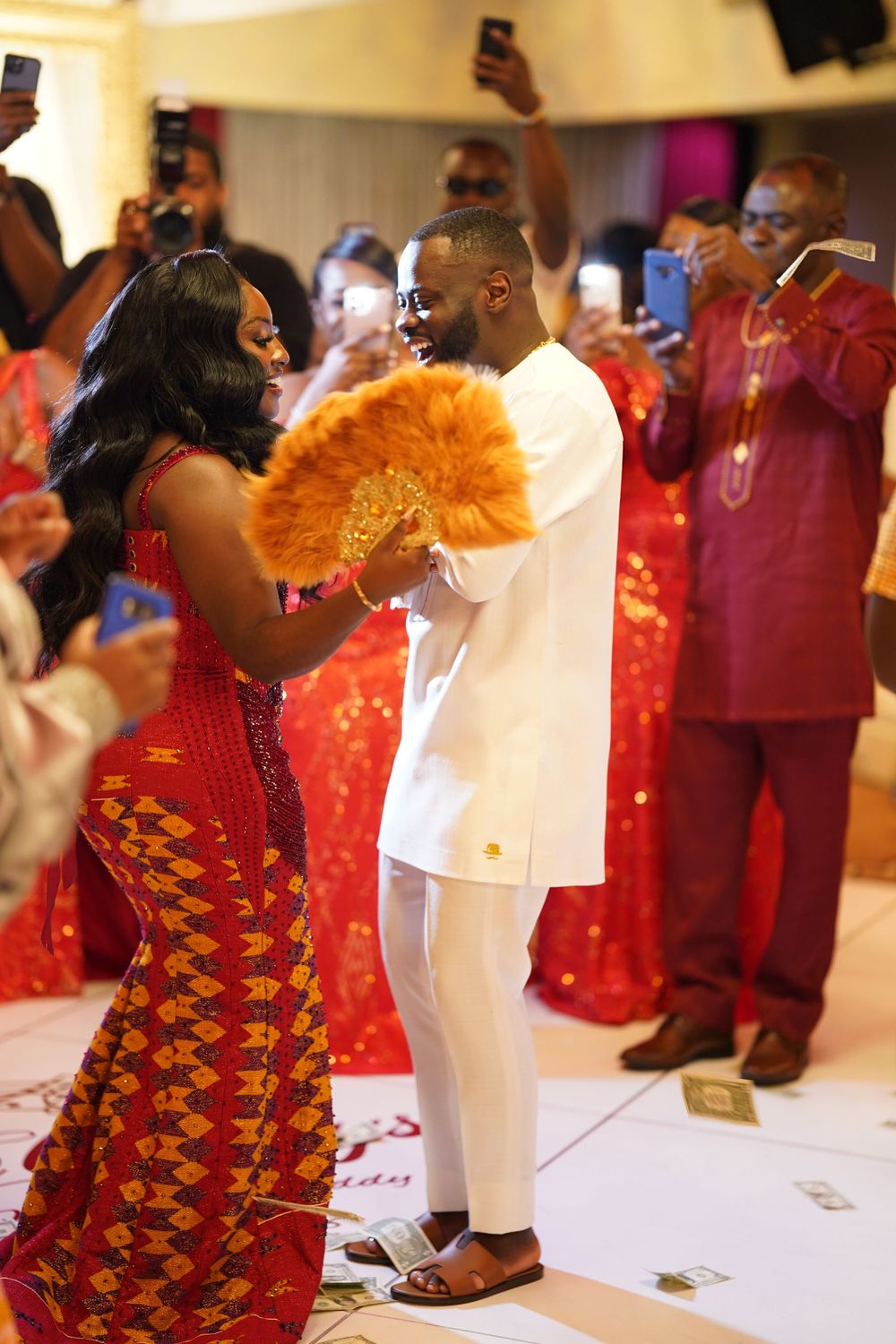 Couple dancing at cultural celebration with guests capturing moment on phones.