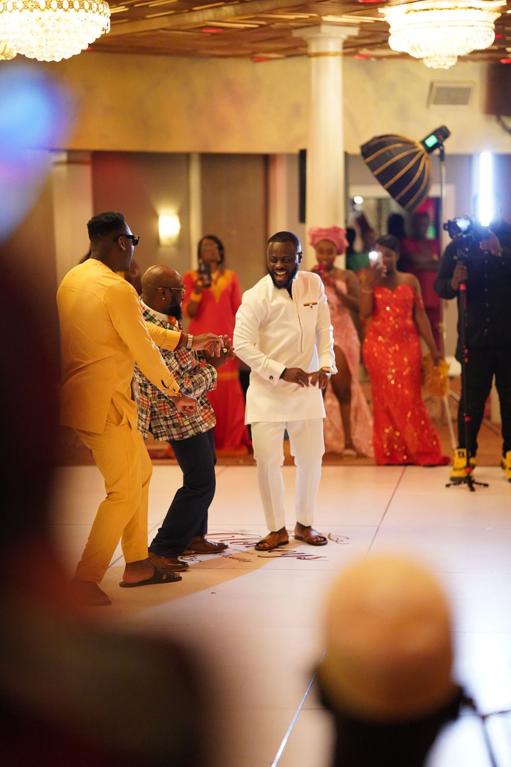 Guests in vibrant formal attire dancing and celebrating at an elegant indoor event with crystal chandeliers.
