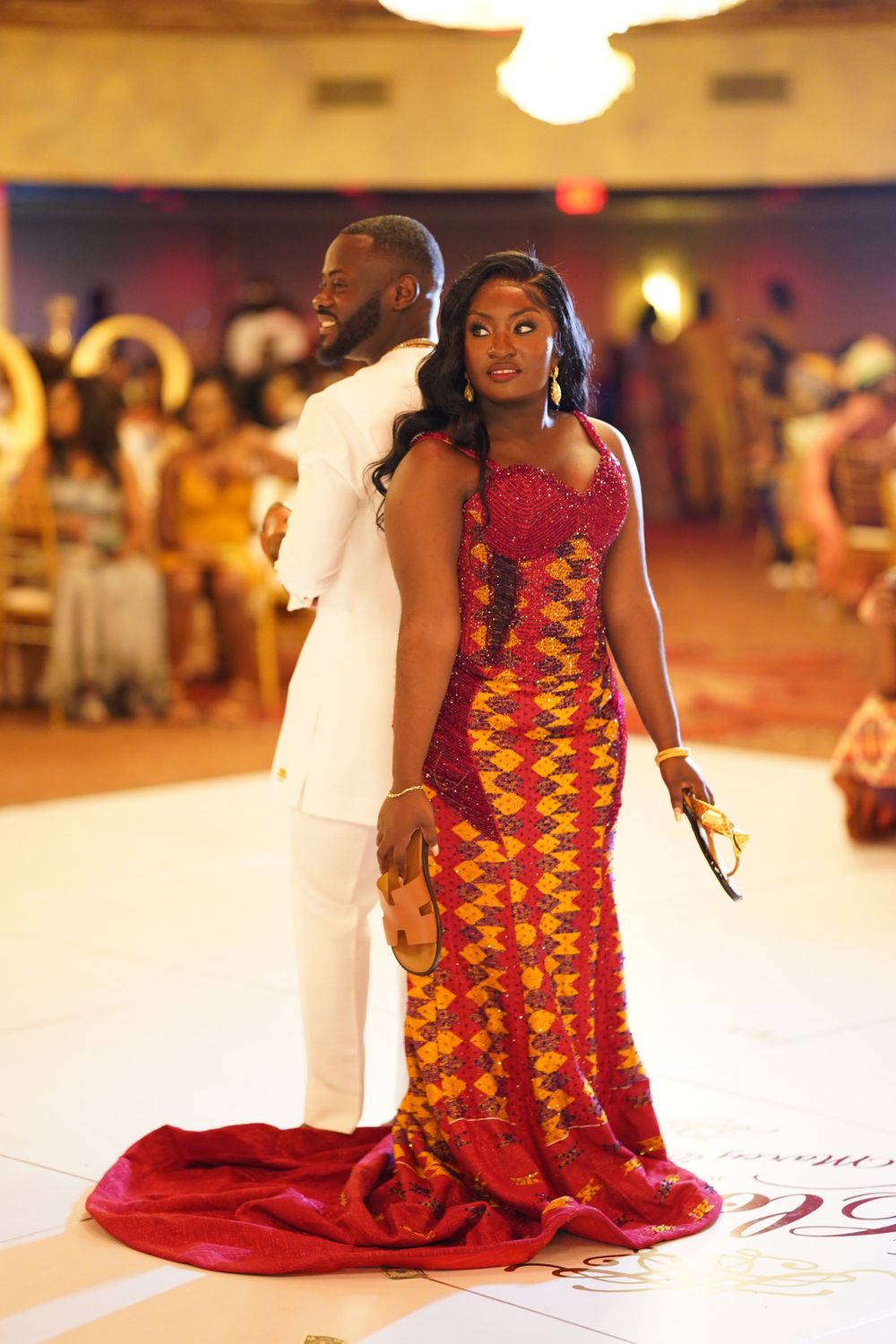 Couple in contrasting white suit and red-yellow African print gown pose during formal celebration.