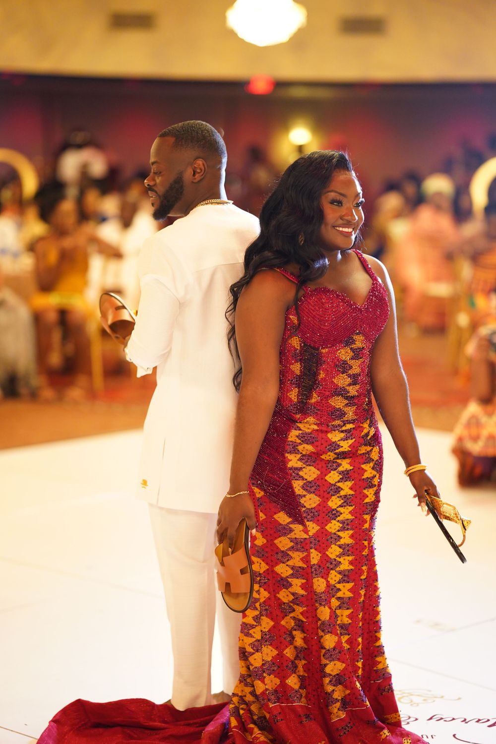 Stylish couple in white suit and patterned African dress stand together at formal celebration.