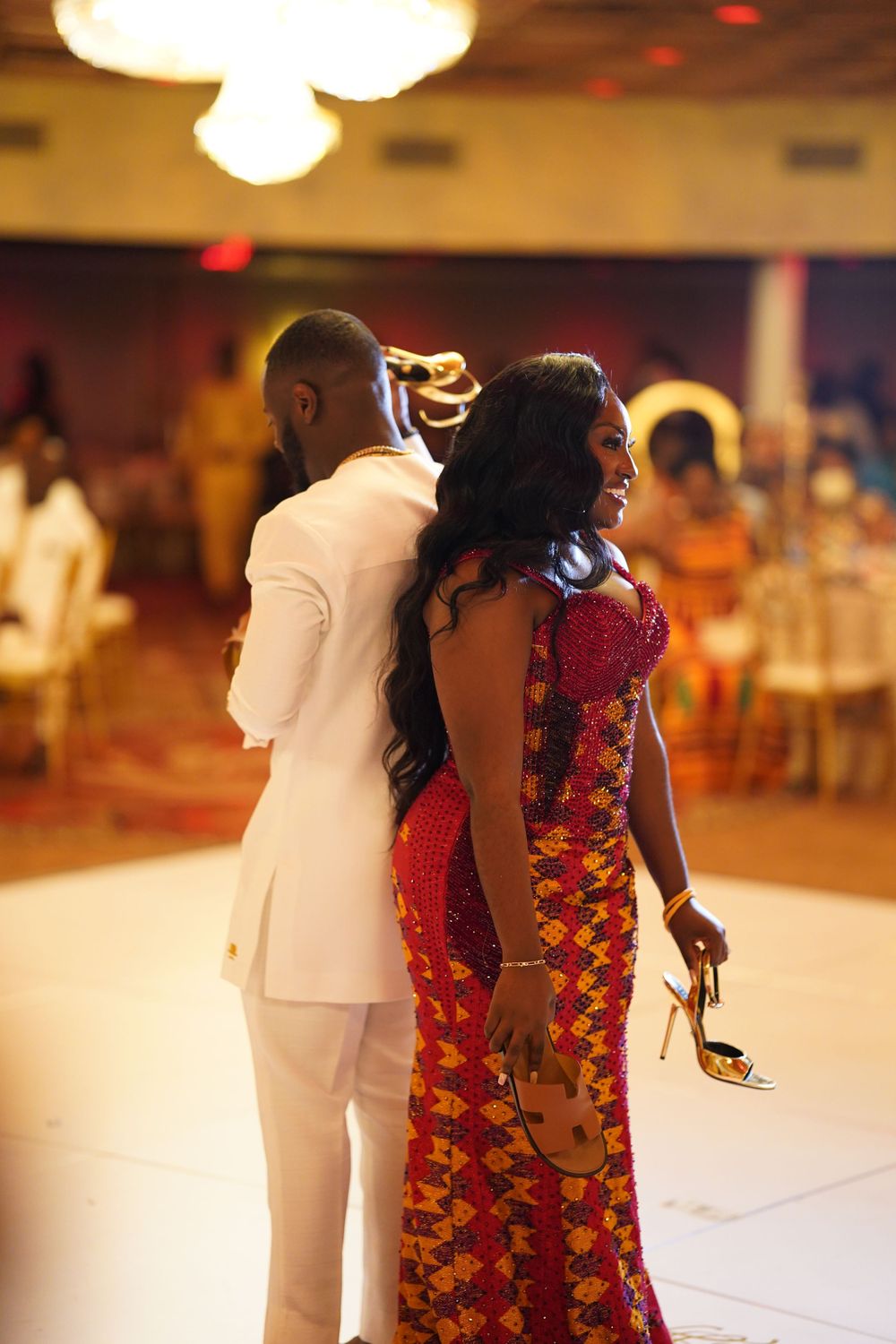 A couple in formal attire dancing at an event, with the woman wearing a vibrant red and gold patterned dress.