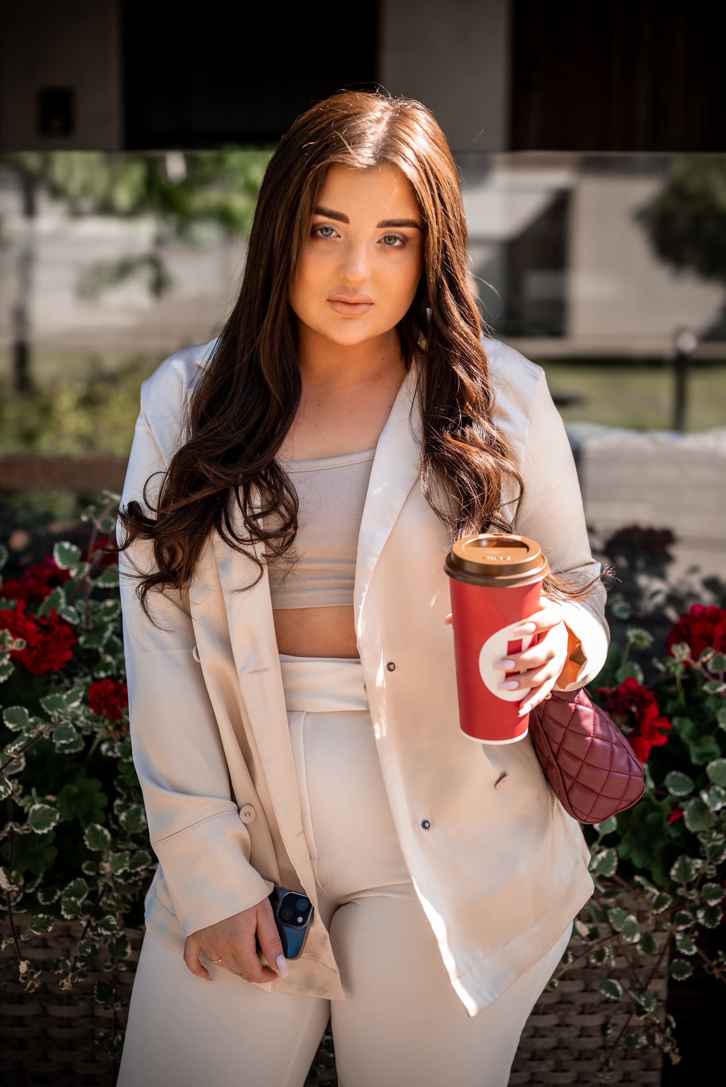 Casual outdoor portrait in cream blazer with red coffee cup near flowers.