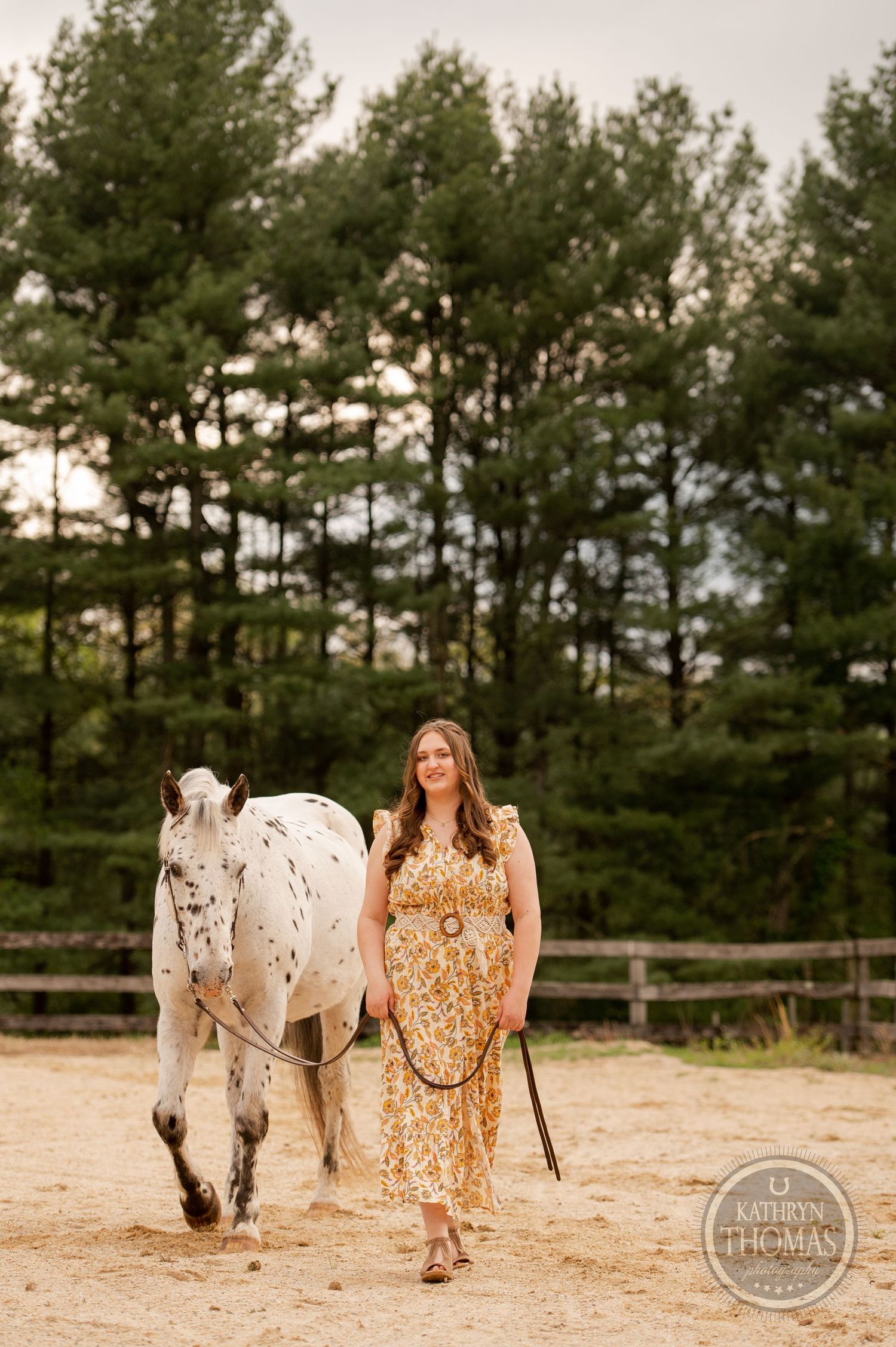 A spotted white horse walks alongside someone in a floral dress near a pine forest fence.