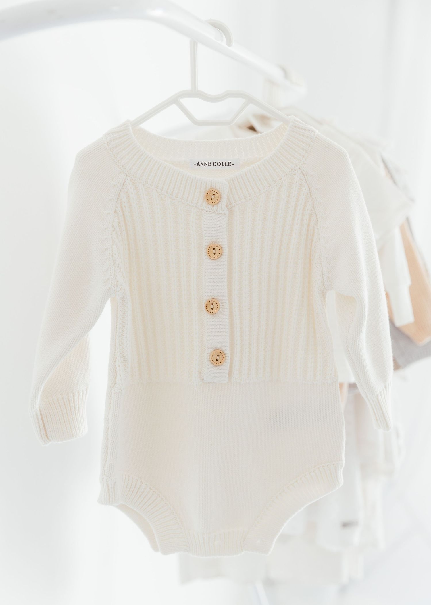 White knitted baby bodysuit with wooden buttons hanging on a clothes hanger.