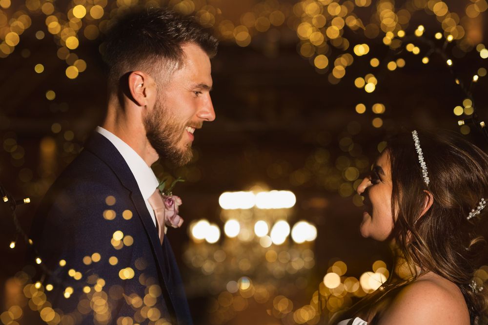 Bride and groom stand face to face in an evening portrait. A blurred chandelier in the background.