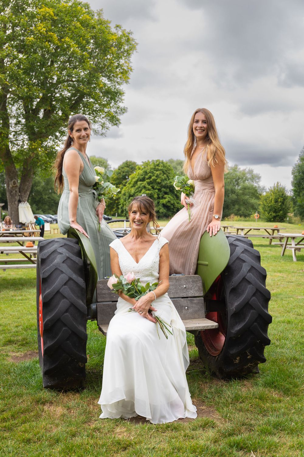 A bride and two bridesmaids pose, sitting on a tractor, with a tree in the background at Arrow Mill.