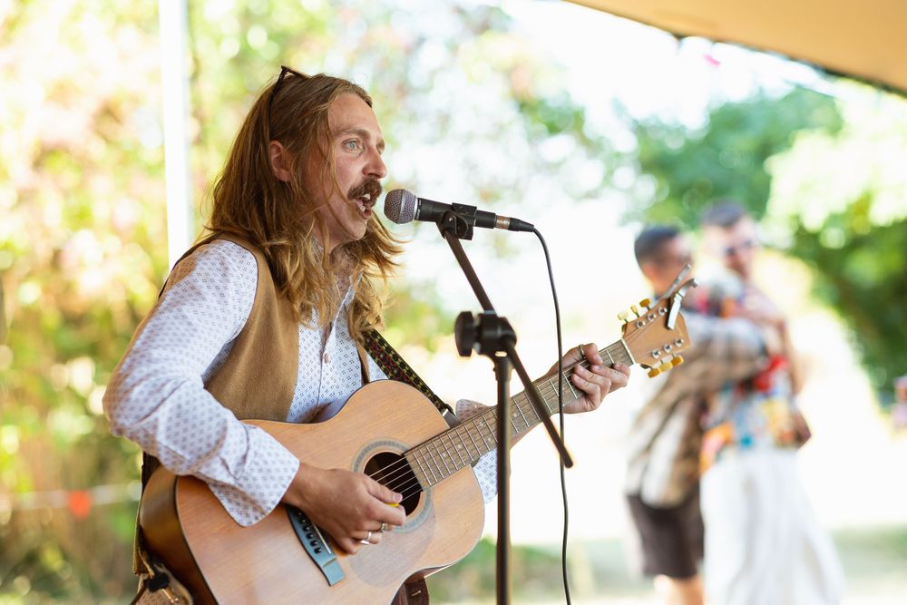 A long-haired man with a moustache and wearing a waistcoat plays an acoustic guitar and sings at a wedding.