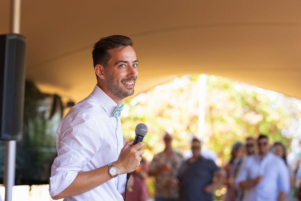 A groom smiles, holding a microphone, as he gives his wedding speech to onlooking wedding guests.