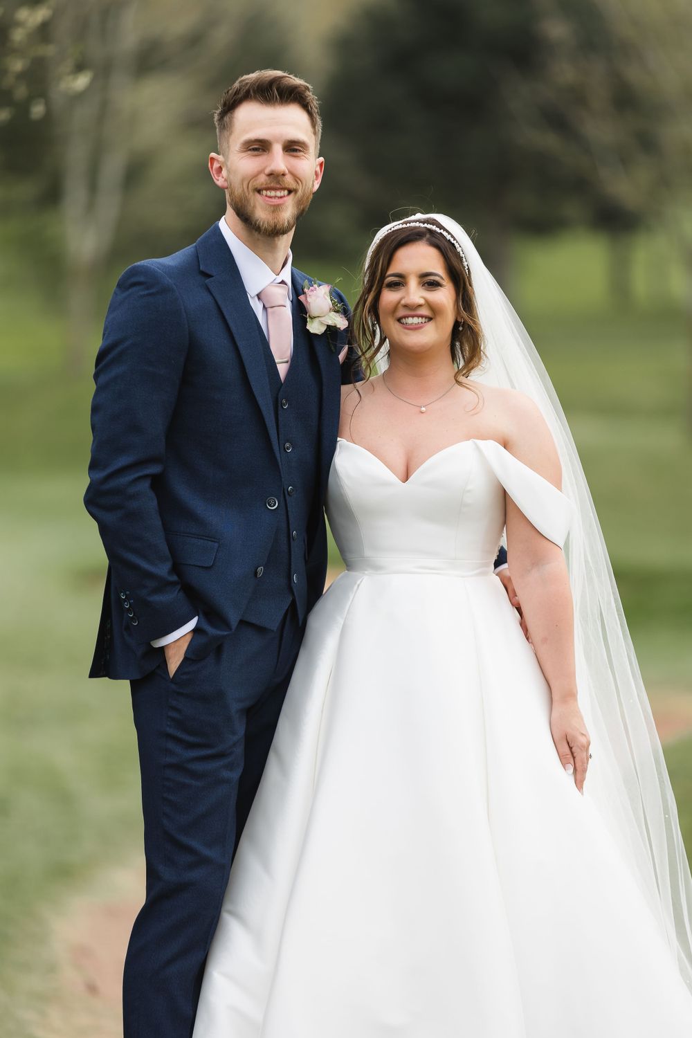 A groom in a navy suits stands next to his bride in a white dress and veil as they look head on to the camera.