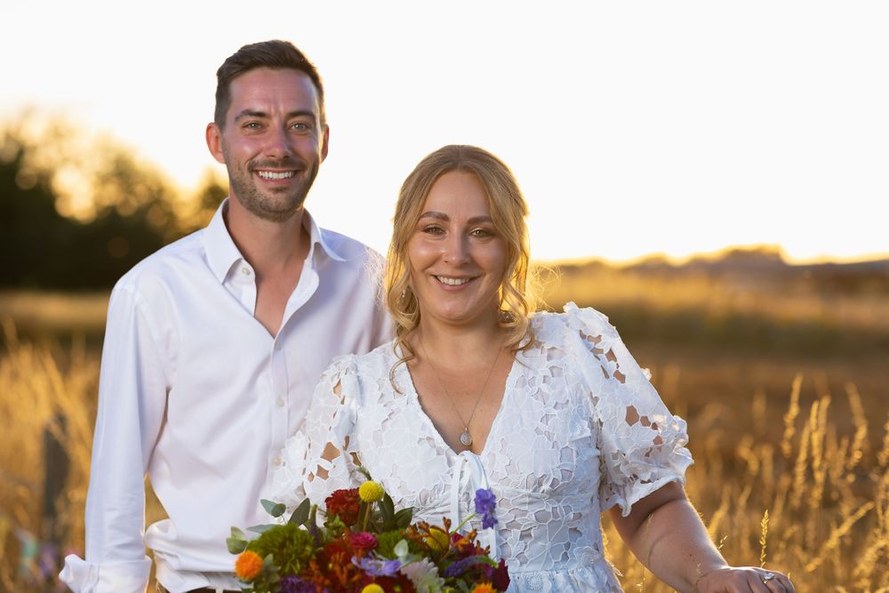 A bride and groom stand facing the camera as the sun sets behind them.