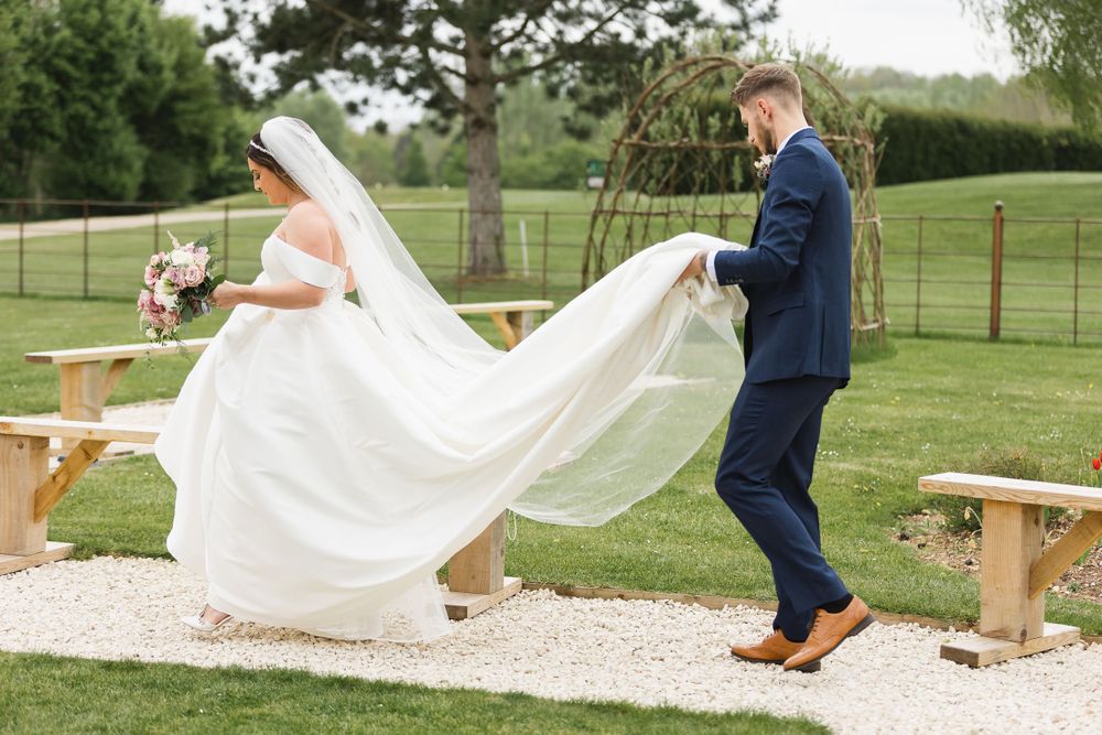 A groom holds up the brides wedding dress as she walks in front of him along a stone path.