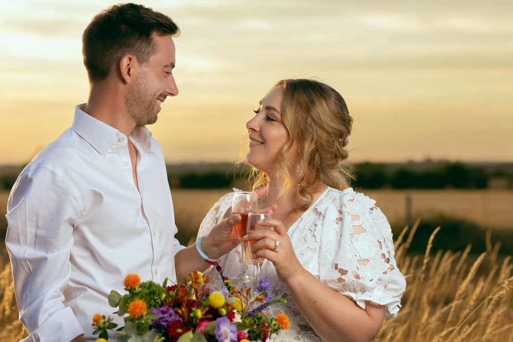 A bride and groom share a toast, clinking glasses of wine together, facing each other while the sun sets behind.