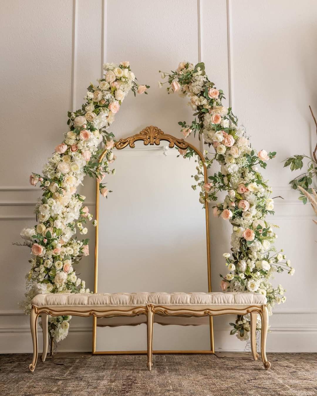 Elegant French-style bench with ornate gold frame and floral arch featuring white and pink roses against a neutral wall.