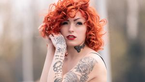 Young woman with curly red hair and tattoos posing outdoors with a soft bokeh background.