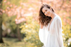 Smiling pregnant woman in white dress surrounded by pink blossoming trees in spring garden.