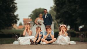 Four children sitting on a path covering their faces while a bride and groom pose romantically in the background.