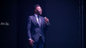 Male performer in grey suit singing passionately into microphone on a dramatic lit stage.