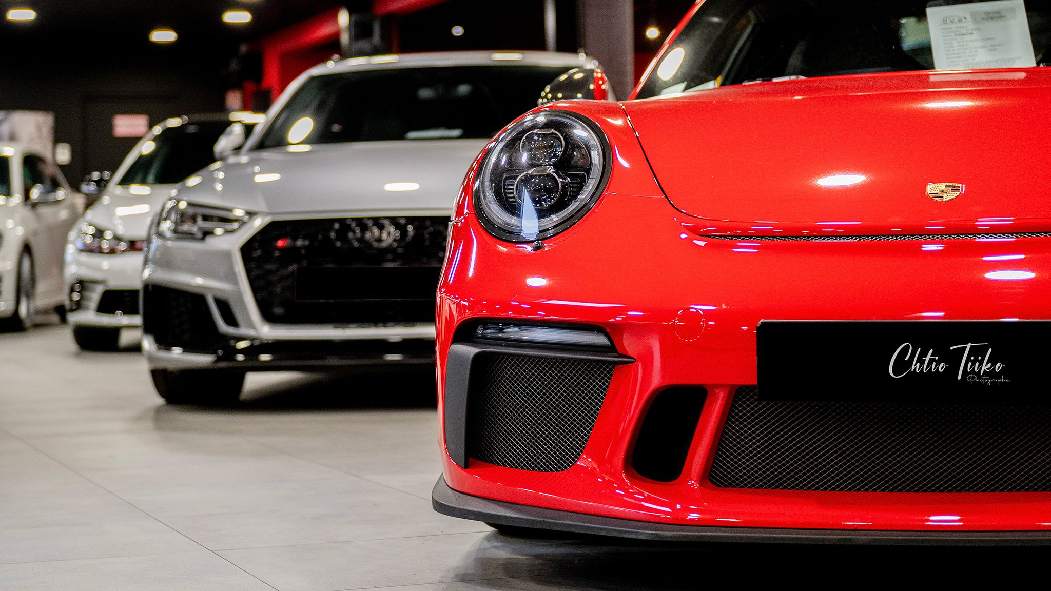 Red Porsche GT3 and silver Audi RS displayed in a modern luxury car dealership showroom.
