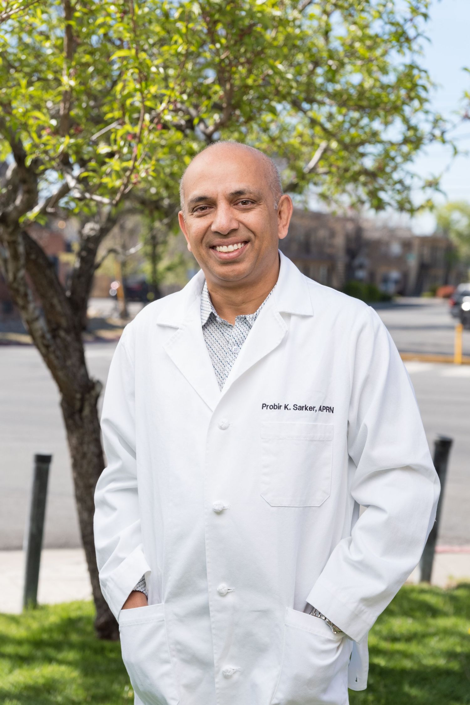Healthcare professional in a white medical coat stands smiling outdoors in front of a leafy tree on a sunny day.
