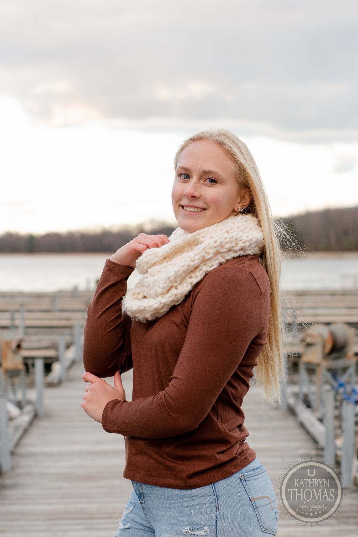 Person in brown sweater and cream scarf smiling on wooden dock with lake view.