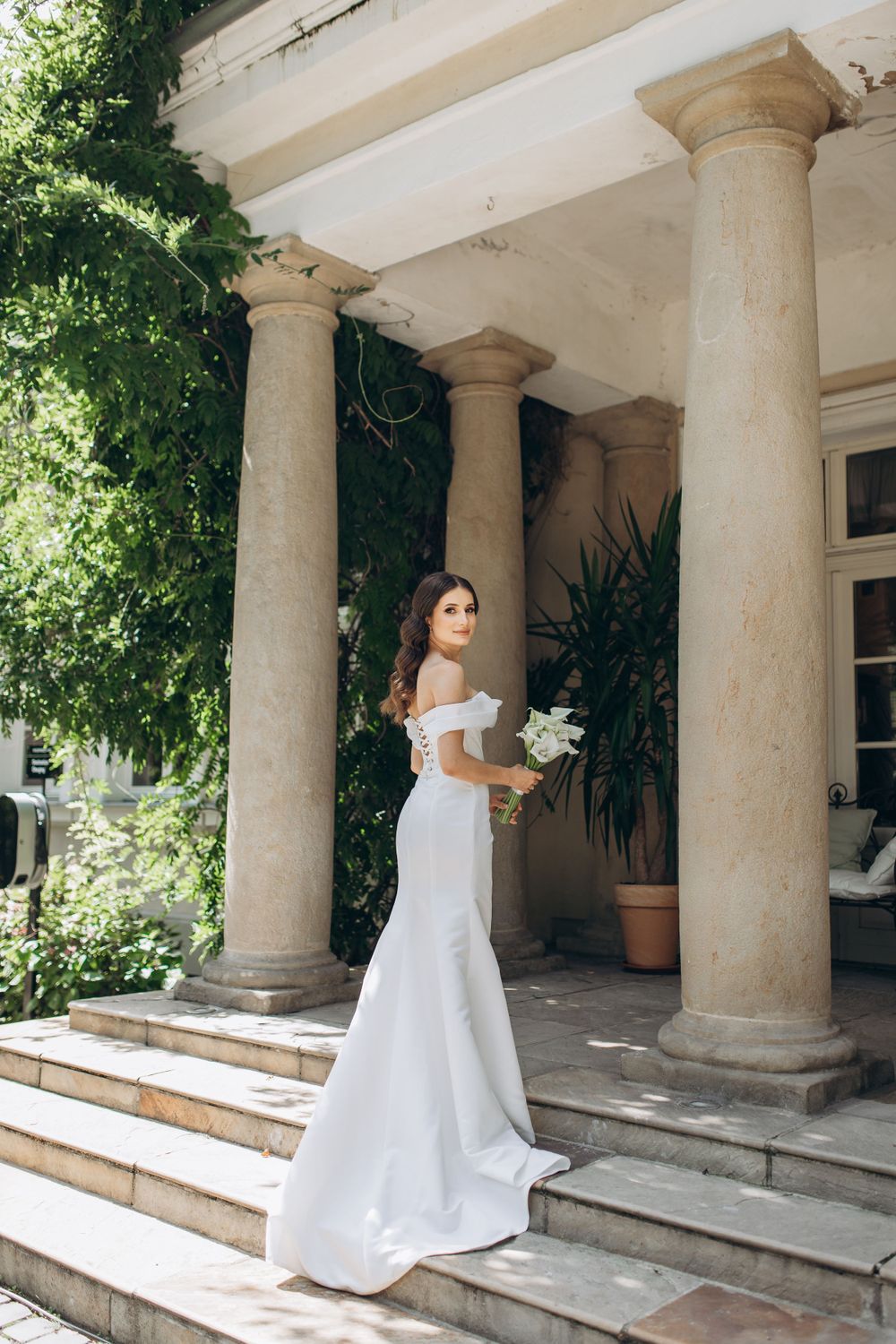 Elegant bridal portrait on stone steps with classical columns and greenery.