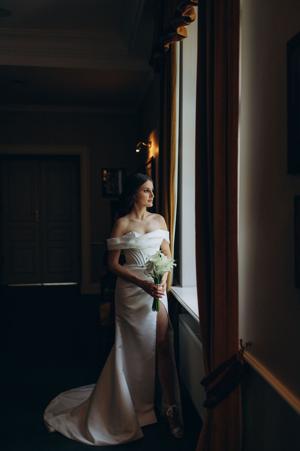 Bride in white dress gazes through window while holding bouquet in moody indoor setting.