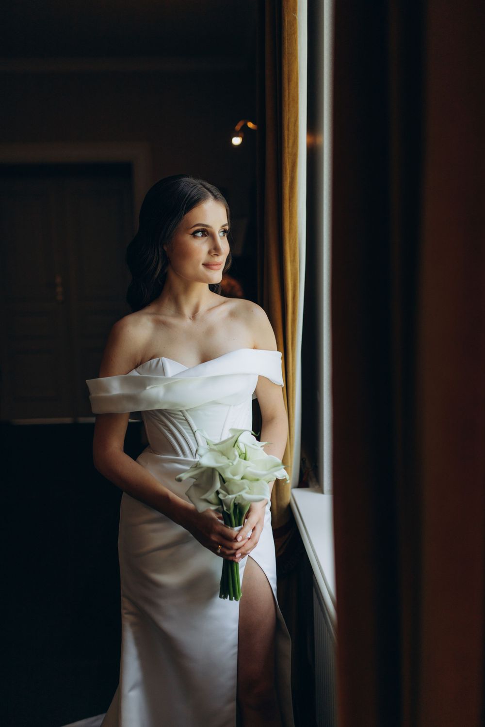 Elegant bride in white dress with thigh slit holds bouquet by window in dramatic lighting.