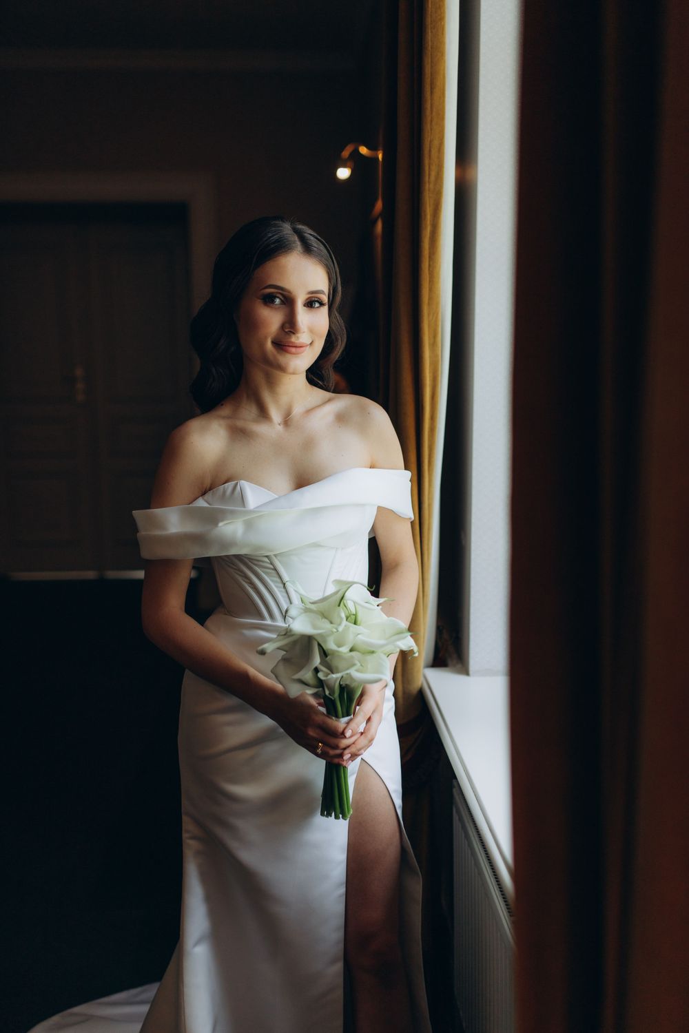 Bride in off-shoulder white gown holds white bouquet while standing by sunlit window.