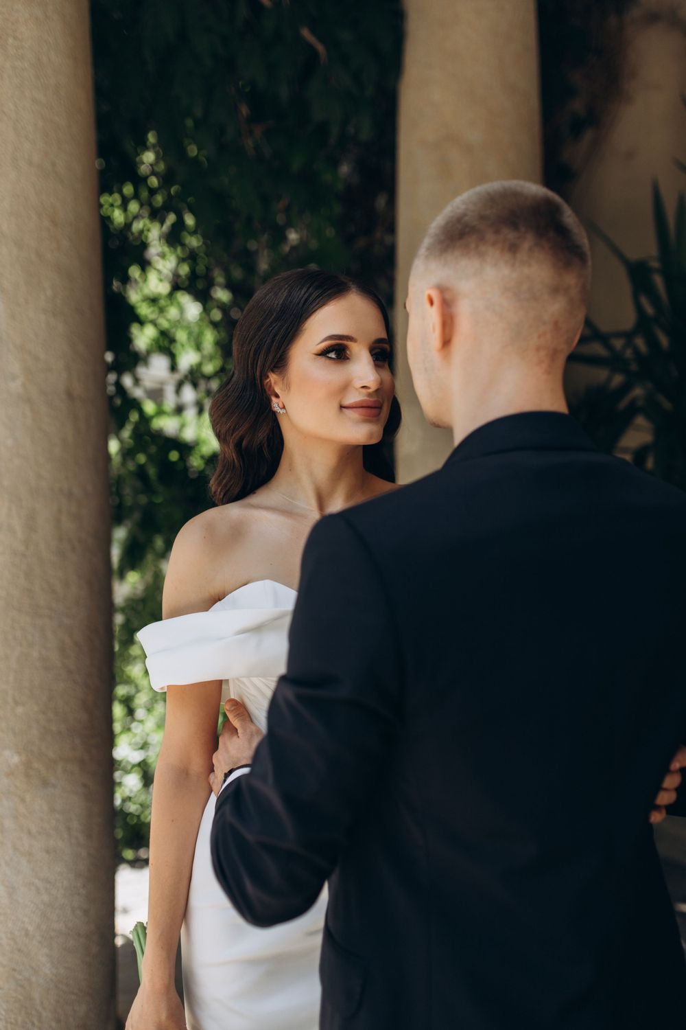 Wedding couple shares intimate moment under stone colonnade with natural greenery.