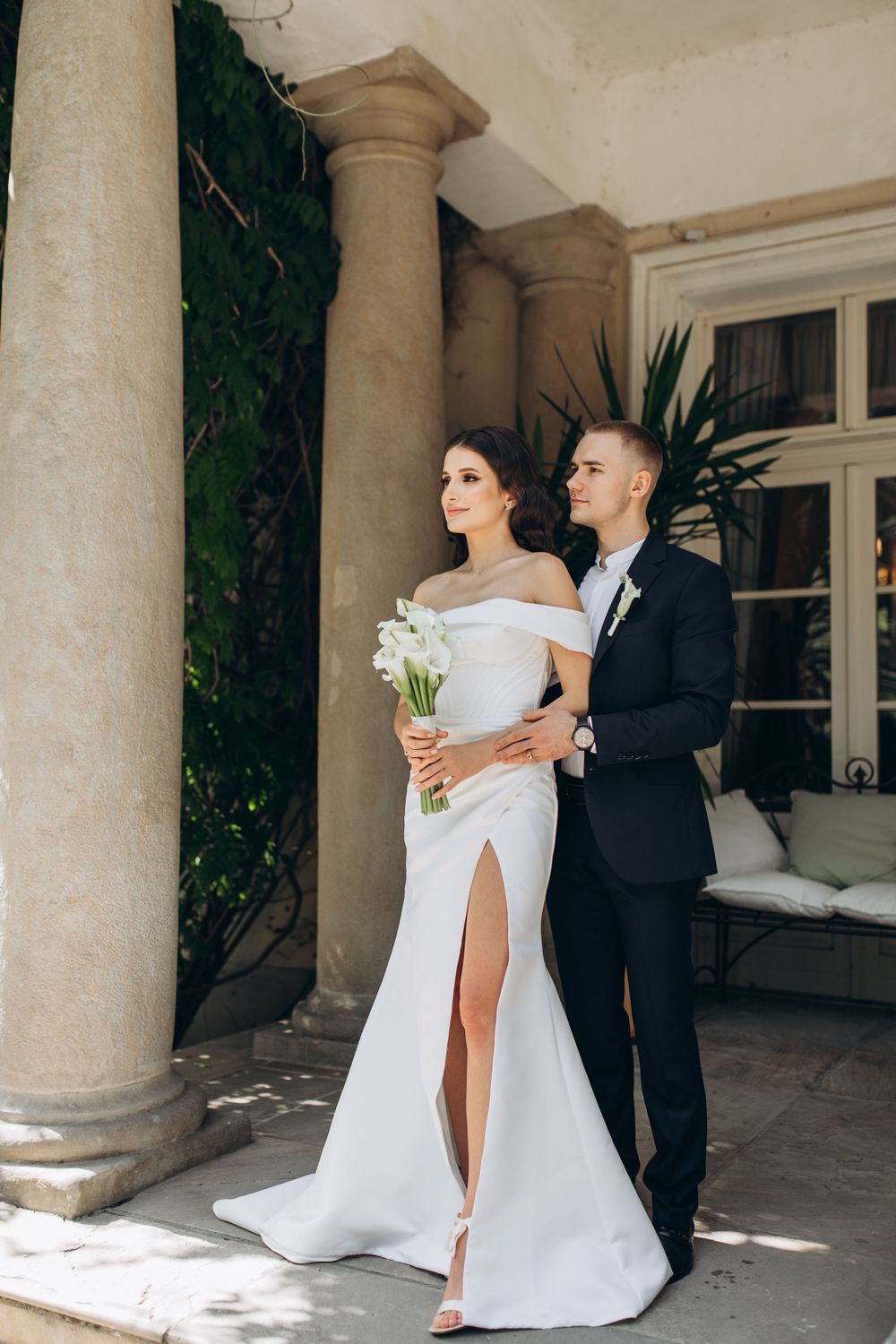Elegant wedding couple posing against stone columns on patio with bride in off-shoulder white gown holding bouquet.