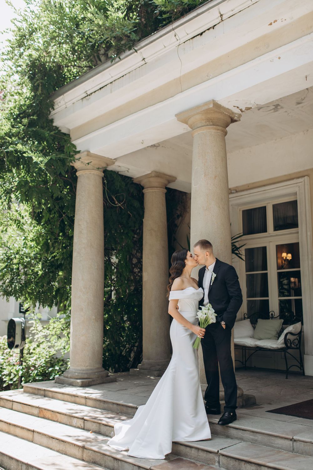 An intimate wedding portrait framed by classic columns on a sun-drenched porch.