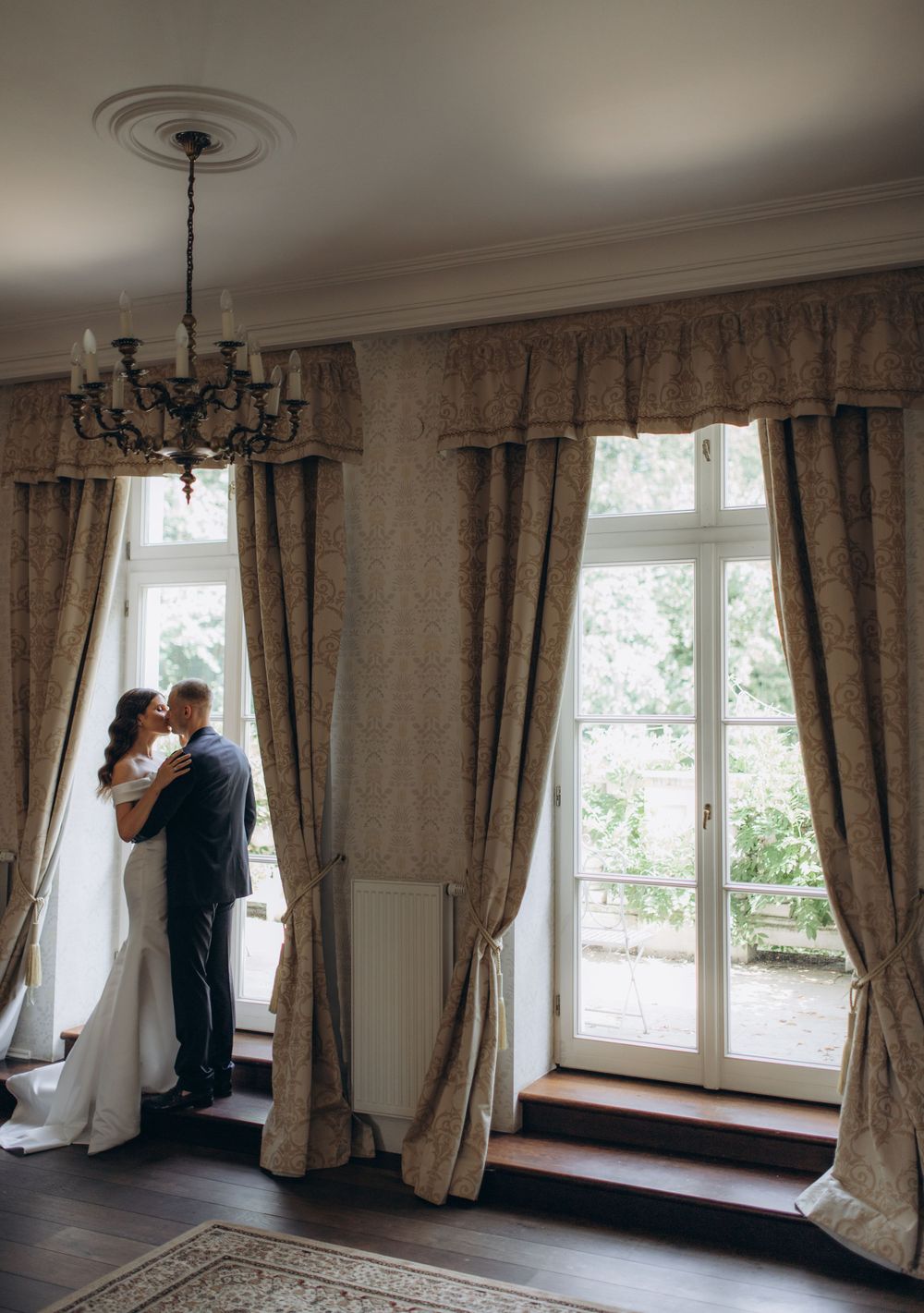 Elegant interior wedding photo with vintage chandelier and floor-length curtains framing the scene.