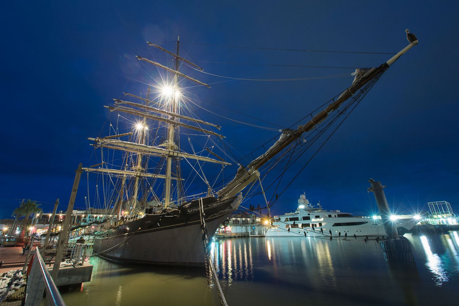 Historic sailing ship illuminated at night in harbor with city lights reflecting on water.