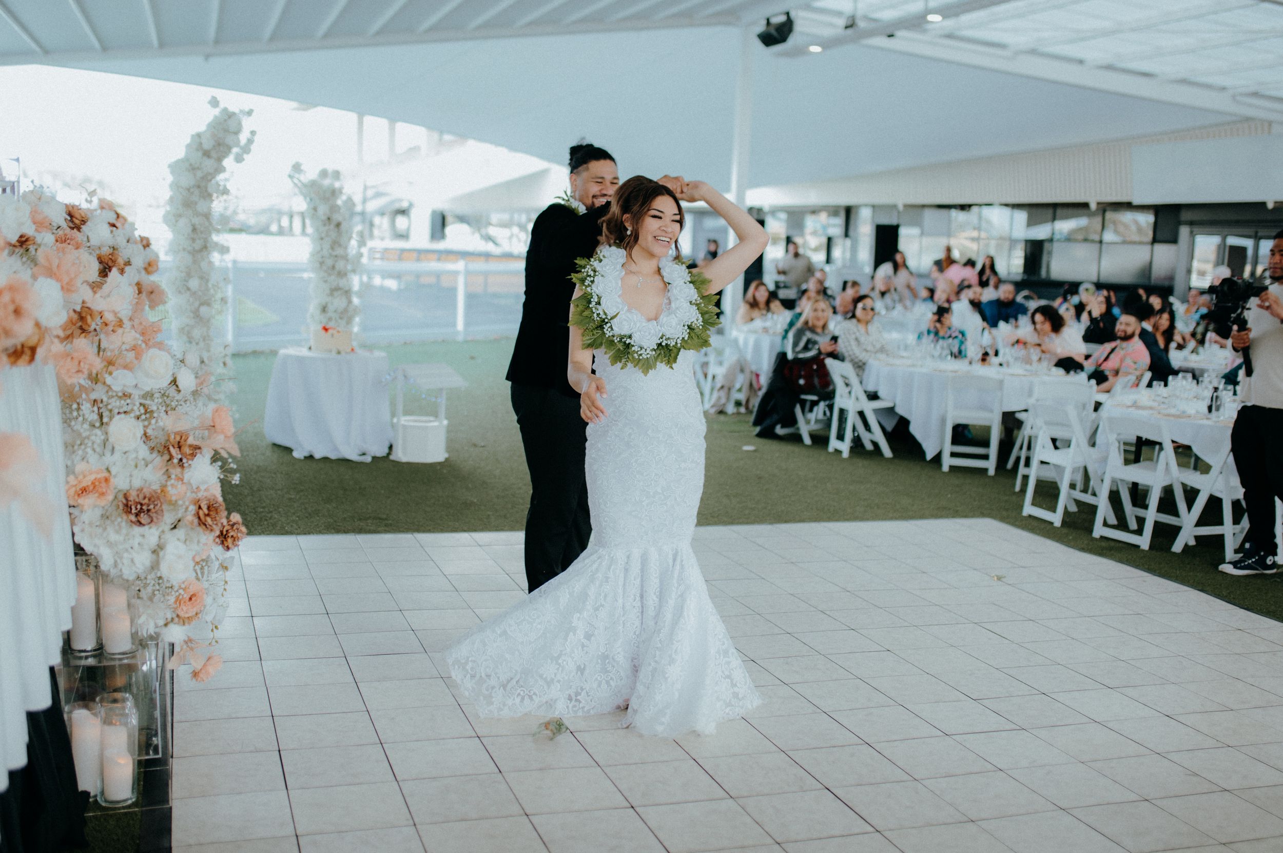 Bride in mermaid wedding gown wearing floral lei entering reception venue with wedding party.