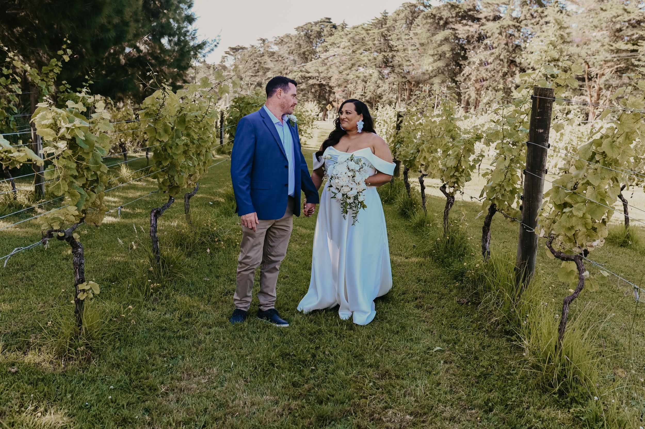 Bride and groom walking through lush green vineyard rows at romantic outdoor winery wedding.
