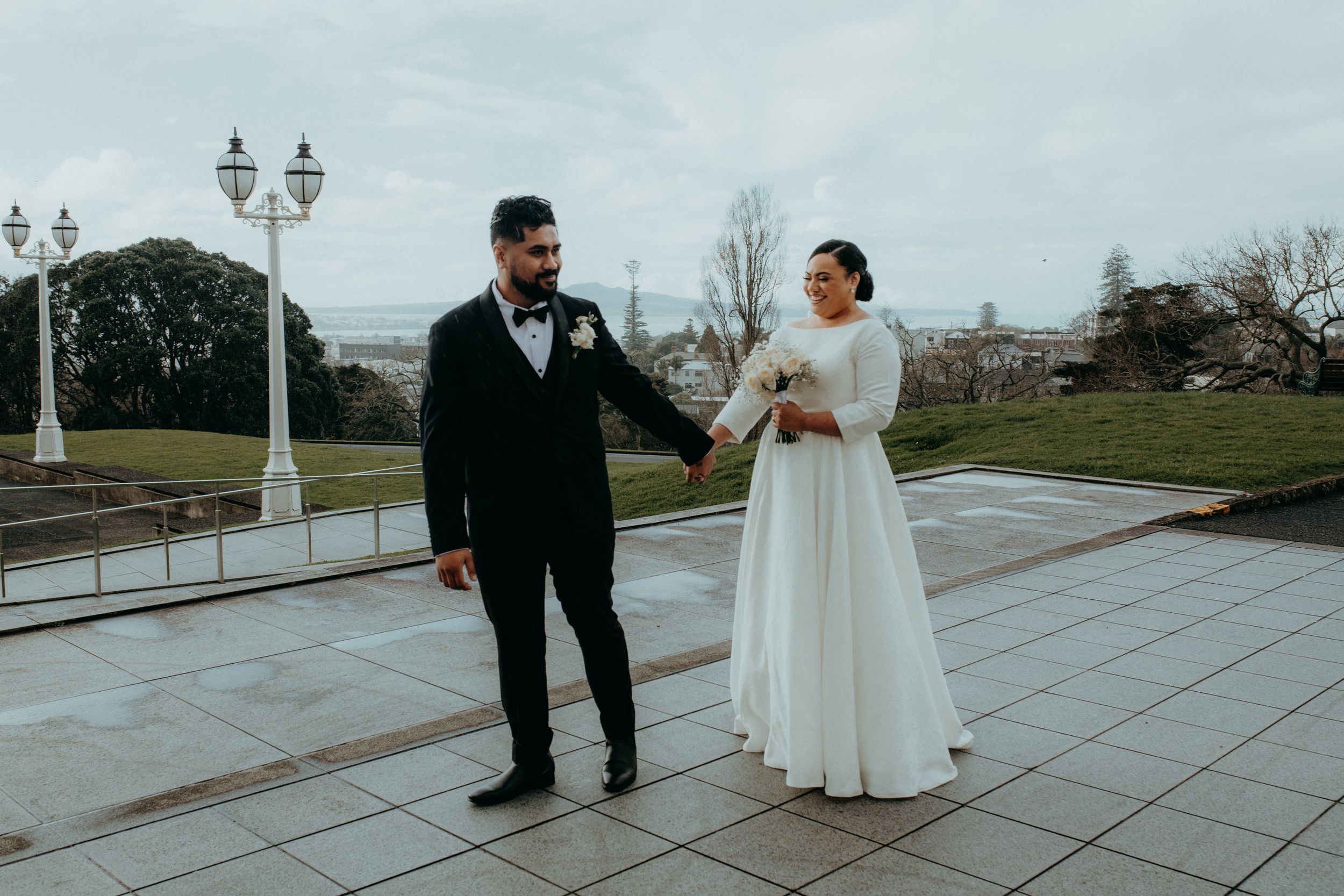 Bride and groom holding hands on a tiled outdoor terrace with green lawns and city views on their wedding day.