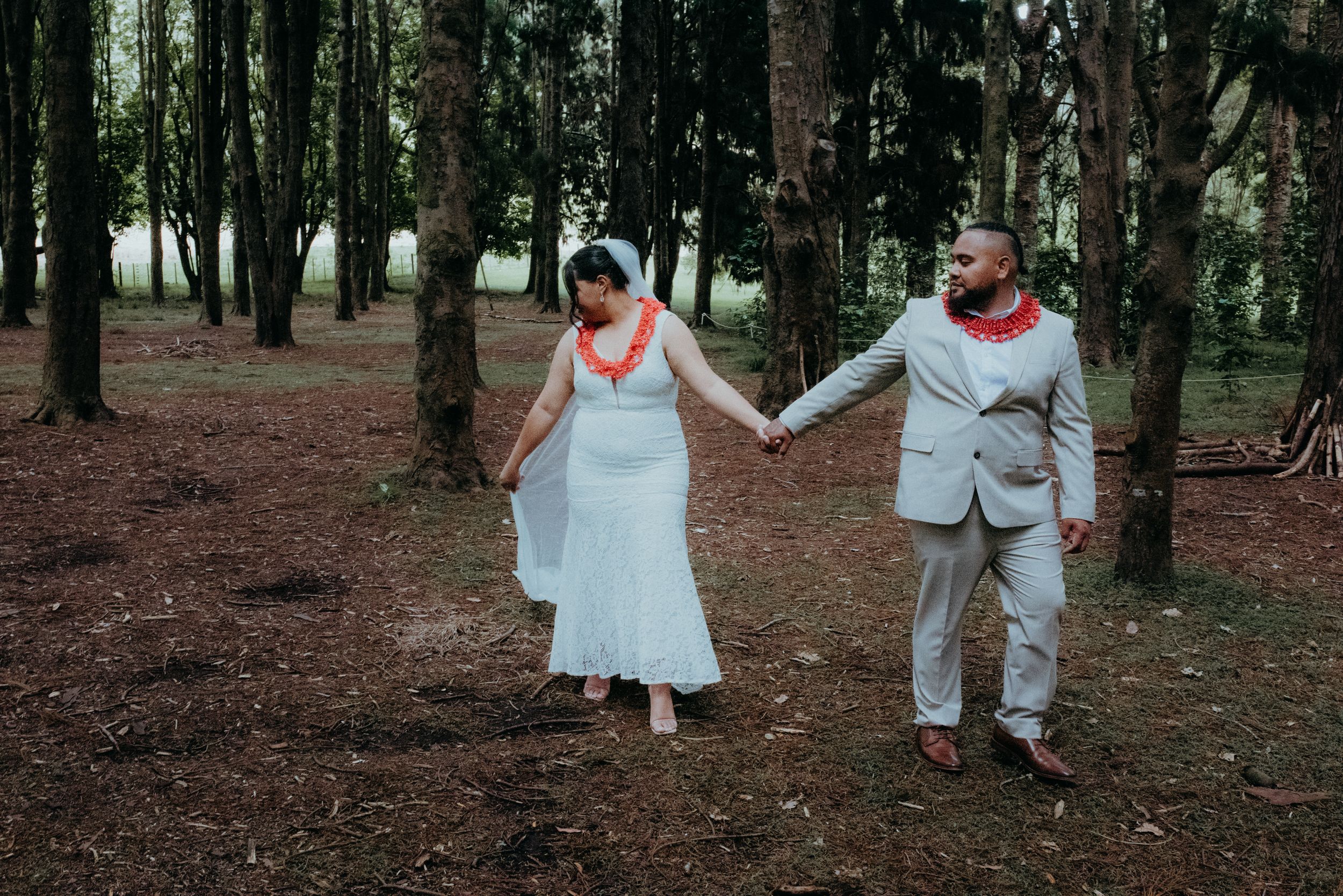 Bride and groom in Polynesian leis hold hands walking through a forest of tall trees.