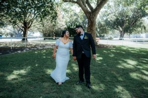 Bride in white gown and groom in black suit holding hands walking in a sunny park.