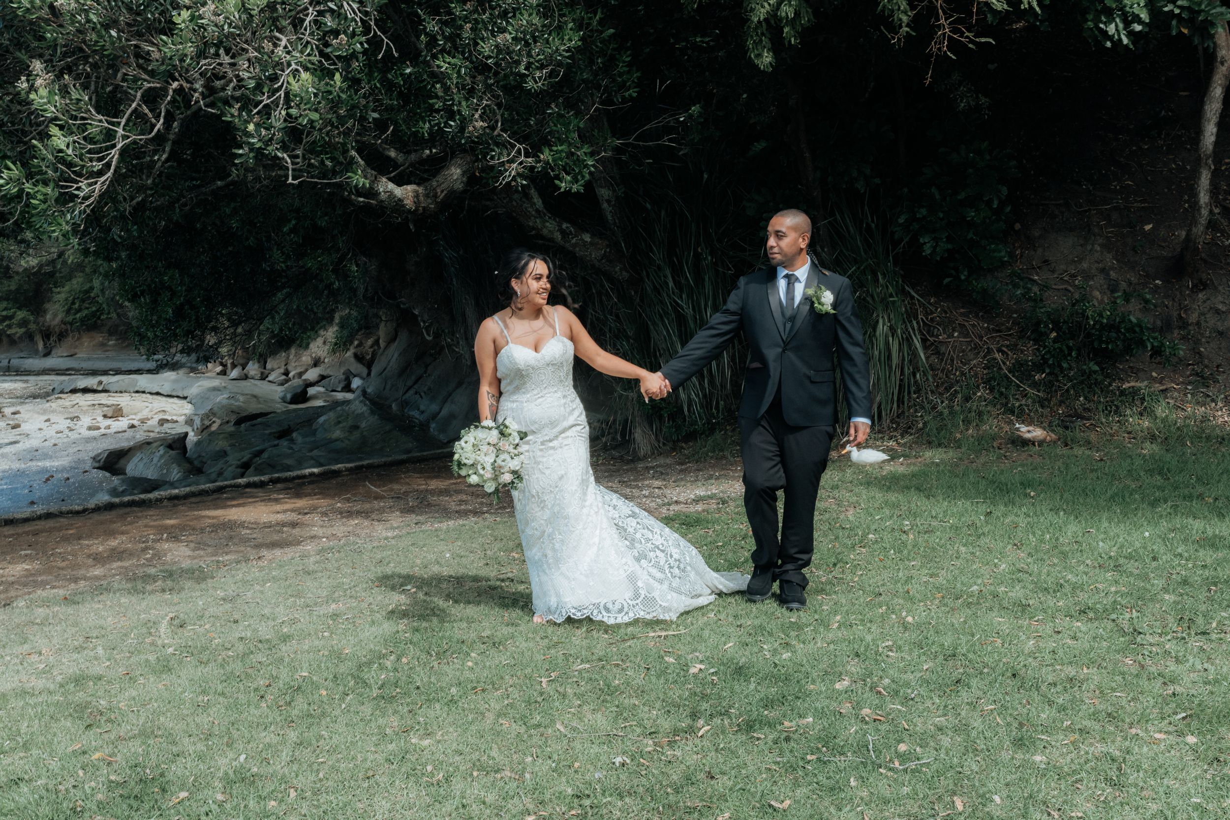 Bride in lace gown and groom in black suit hold hands walking on grass near trees.