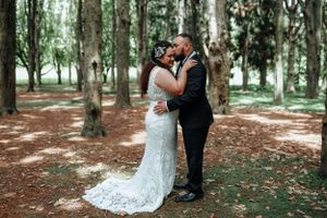Bride and groom share a passionate kiss among tall trees in a sunlit forest.
