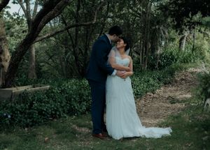 Newlyweds share a romantic kiss surrounded by lush green trees in outdoor garden setting.