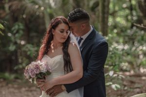 Groom tenderly embraces bride holding pink bouquet in a lush forest wedding portrait.
