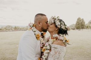Couple sharing a romantic kiss wearing floral leis and flower crown at an outdoor wedding ceremony.